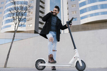 Handsome man with electric scooter talking on smart phone while looking away against buildings - EGAF01700