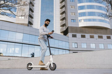 Young man with electric scooter using smart phone while standing against buildings in city during COVID-19 - EGAF01701