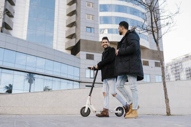 Mid adult man with electric scooter smiling at male friend while walking through city - EGAF01703