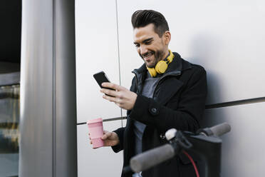 Lächelnder Mann mit Kaffeetasse, der sein Smartphone benutzt, während er mit einem Elektroroller an der Wand steht - EGAF01713