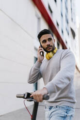 Junger Mann, der mit seinem Smartphone spricht, während er neben einem elektrischen Roller steht und gegen ein Gebäude schaut - EGAF01721