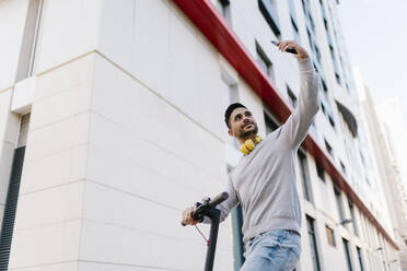Junger Mann, der ein Selfie mit seinem Smartphone macht, während er mit einem Elektroroller vor einem Gebäude steht - EGAF01722