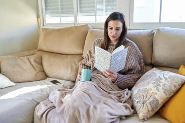 Frau liest ein Buch und hält eine Tasse auf dem Sofa im Winter - KIJF03579