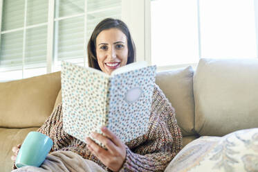 Lächelnde Frau mit Tasse und Buch auf dem Sofa zu Hause - KIJF03581