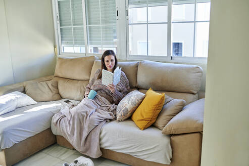 Frau mit Tasse, die ein Buch liest, während sie zu Hause auf dem Sofa sitzt - KIJF03582