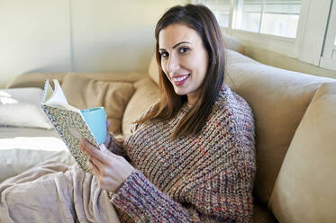 Lächelnde schöne Frau mit Tasse hält Buch auf Sofa zu Hause - KIJF03583