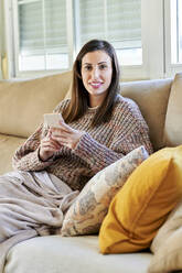Lächelnde Frau, die ihr Handy hält, während sie zu Hause auf dem Sofa sitzt - KIJF03586