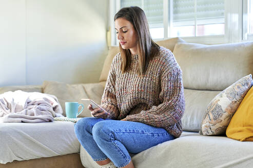Frau in warmer Kleidung mit Smartphone auf dem Sofa zu Hause - KIJF03587