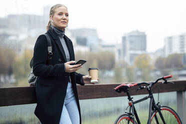 Woman holding mobile phone while listening music through headphones by bicycle - JSRF01383