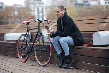 Frau benutzt Mobiltelefon, während sie auf einer Bank am Fahrrad sitzt - JSRF01385