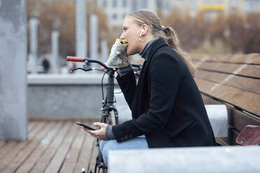 Frau mit Smartphone isst Wrap-Sandwich, während sie auf einer Bank am Fahrrad sitzt - JSRF01387