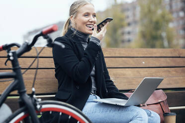 Lachende Frau mit Laptop, die mit ihrem Handy telefoniert, während sie auf einer Bank am Fahrrad sitzt - JSRF01388