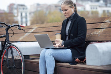 Frau hält Kaffeetasse, während sie einen Laptop auf einer Bank am Fahrrad benutzt - JSRF01389