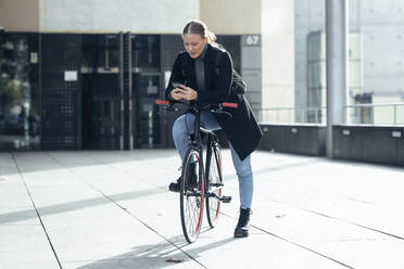 Junge Frau benutzt ein Mobiltelefon auf einem Fahrrad vor einem Gebäude - JSRF01391