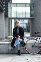 Woman sitting on retaining wall against structure by bicycle - JSRF01393