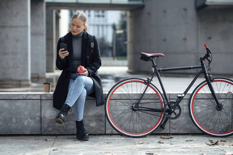 Young woman using mobile phone while sitting on retaining wall by bicycle stock photo