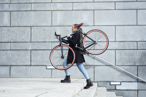 Frau, die ein Fahrrad trägt, während sie auf einer Treppe an der Wand läuft - JSRF01404