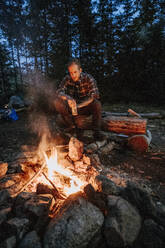Junger Mann in Flanell mit Bier in der Hand sitzt nachts am Lagerfeuer in Maine - CAVF93053