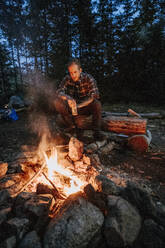 Young man in flannel holding beer sits by campfire at night in Maine - CAVF93053