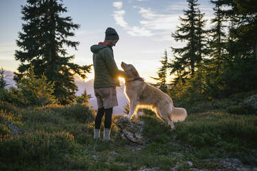 Männlicher Wanderer streichelt Hund bei Sonnenuntergang in den Bergen - CAVF93087