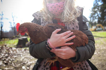 Nahaufnahme eines Mädchens mit einer Maske, das ein Huhn auf einem Bauernhof hält - CAVF93101