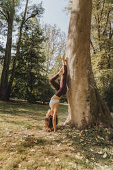 Sportswoman doing handstand yoga while leaning on tree at park - MFF07098