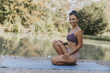 Female athlete smiling while sitting on exercise mat by lake at park - MFF07111