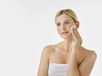 Woman wrapped in towel using cotton pad while standing white background - RORF02584