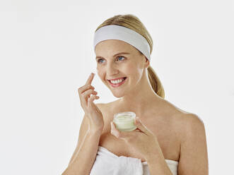 Young woman smiling while applying facial cream standing against white background - RORF02590