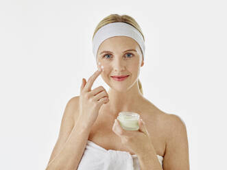 Woman wearing headband and towel applying face cream while standing against white background - RORF02591
