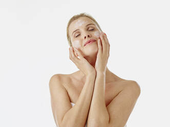 Woman wrapped in towel applying face cream while standing against white background - RORF02597