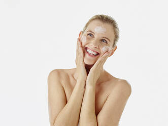 Smiling woman rubbing cream on face while standing against white background - RORF02599