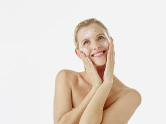 Young woman smiling while applying face cream standing against white background - RORF02600