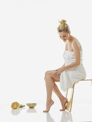Frau in Handtuch Anwendung Feuchtigkeitscreme auf Bein, während auf Stuhl gegen weißen Hintergrund sitzen - RORF02605
