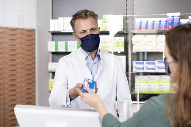 Man in protective mask giving medicine to female customer at pharmacy checkout - FKF03962