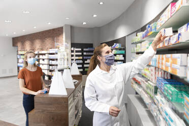 Customer standing at checkout looking at female pharmacist taking medicine from shelf in store - FKF03975