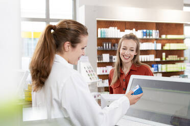 Female pharmacist reading instructions on medicine while woman standing at checkout - FKF03989