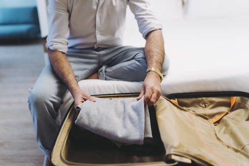 Man unpacking luggage while sitting on bed in hotel room - DGOF01909