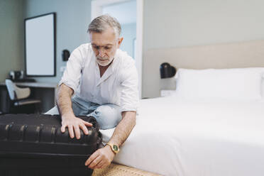 Man closing suitcase while sitting on bed in hotel room - DGOF01910