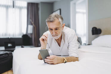 Mann benutzt sein Smartphone, während er auf dem Bett im Hotelzimmer liegt - DGOF01913