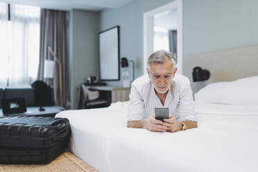 Mann mit Brille liegt auf dem Bett und benutzt sein Smartphone im Hotelzimmer - DGOF01914