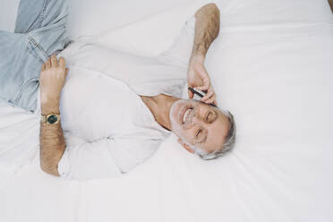 Lächelnder Mann, der auf dem Bett liegend mit seinem Smartphone spricht - DGOF01915