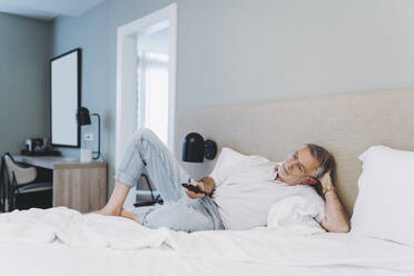 Entspannter Mann, der die Fernbedienung benutzt, während er auf dem Bett im Hotelzimmer liegt - DGOF01917