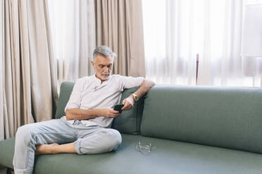 Mann benutzt sein Smartphone, während er auf dem Sofa vor den Vorhängen im Hotelzimmer sitzt - DGOF01925