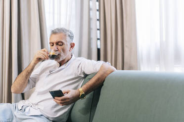 Älterer Mann, der Kaffee trinkt und sein Smartphone benutzt, während er auf dem Sofa im Hotelzimmer sitzt - DGOF01926