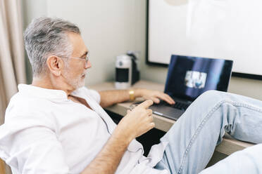 Mature man using laptop with feet up at desk while having coffee in hotel room - DGOF01928