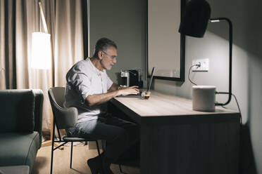 Älterer Mann sitzt mit Kaffee am Schreibtisch und benutzt einen Laptop im Hotelzimmer - DGOF01929