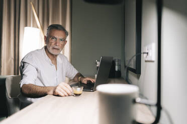 Reifer Mann hält Kaffeetasse, während er am Schreibtisch mit Laptop in einem Hotelzimmer sitzt - DGOF01931