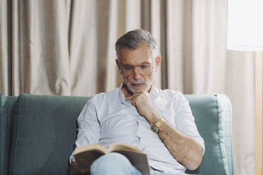 Thoughtful man with hand on chin reading book while sitting on sofa in hotel room - DGOF01932