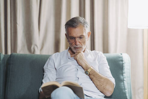 Nachdenklicher Mann mit Hand am Kinn, der ein Buch liest, während er auf dem Sofa im Hotelzimmer sitzt - DGOF01932